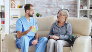 Male nurse in blue scrubs sitting on a couch with an elderly woman, offering her a glass of juice. Home healthcare support in Philadelphia.